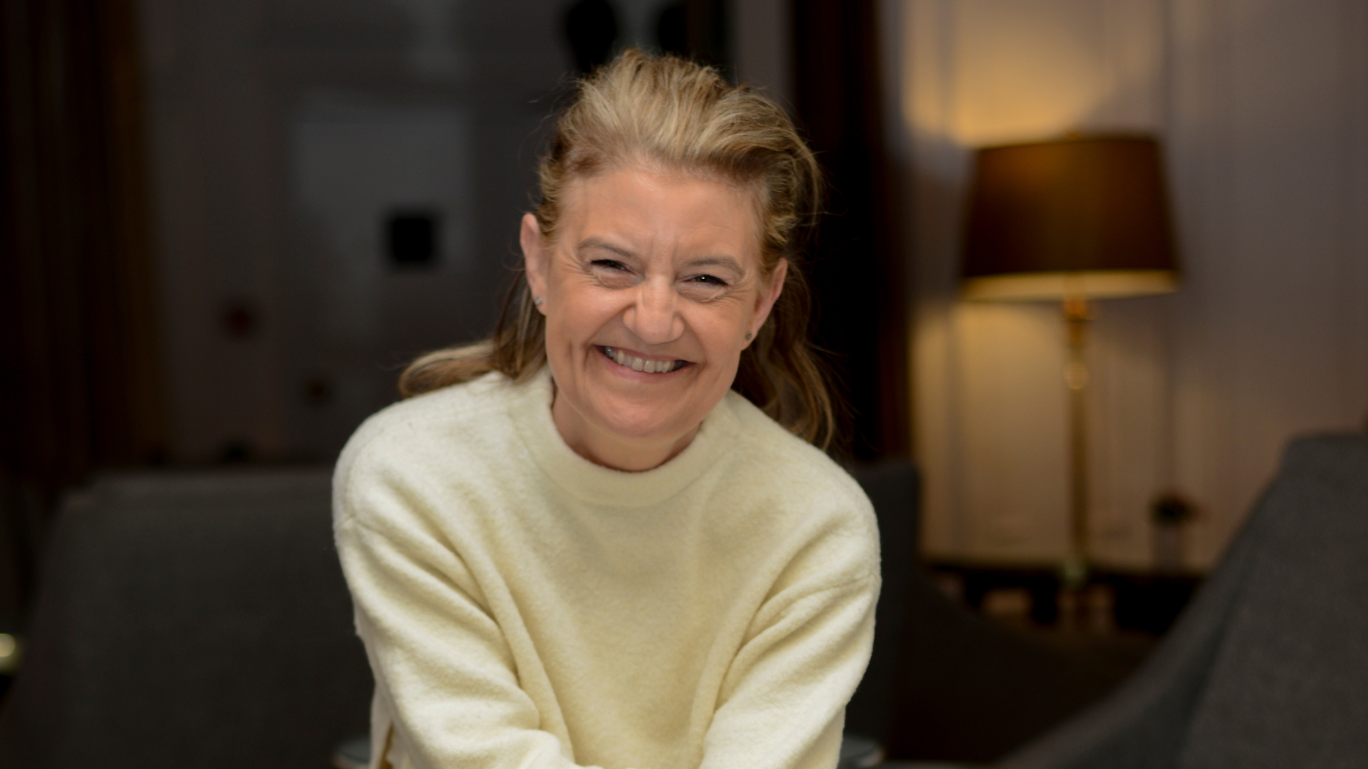 Smiling woman in a cream sweater sitting indoors with a warm lamp in the background.