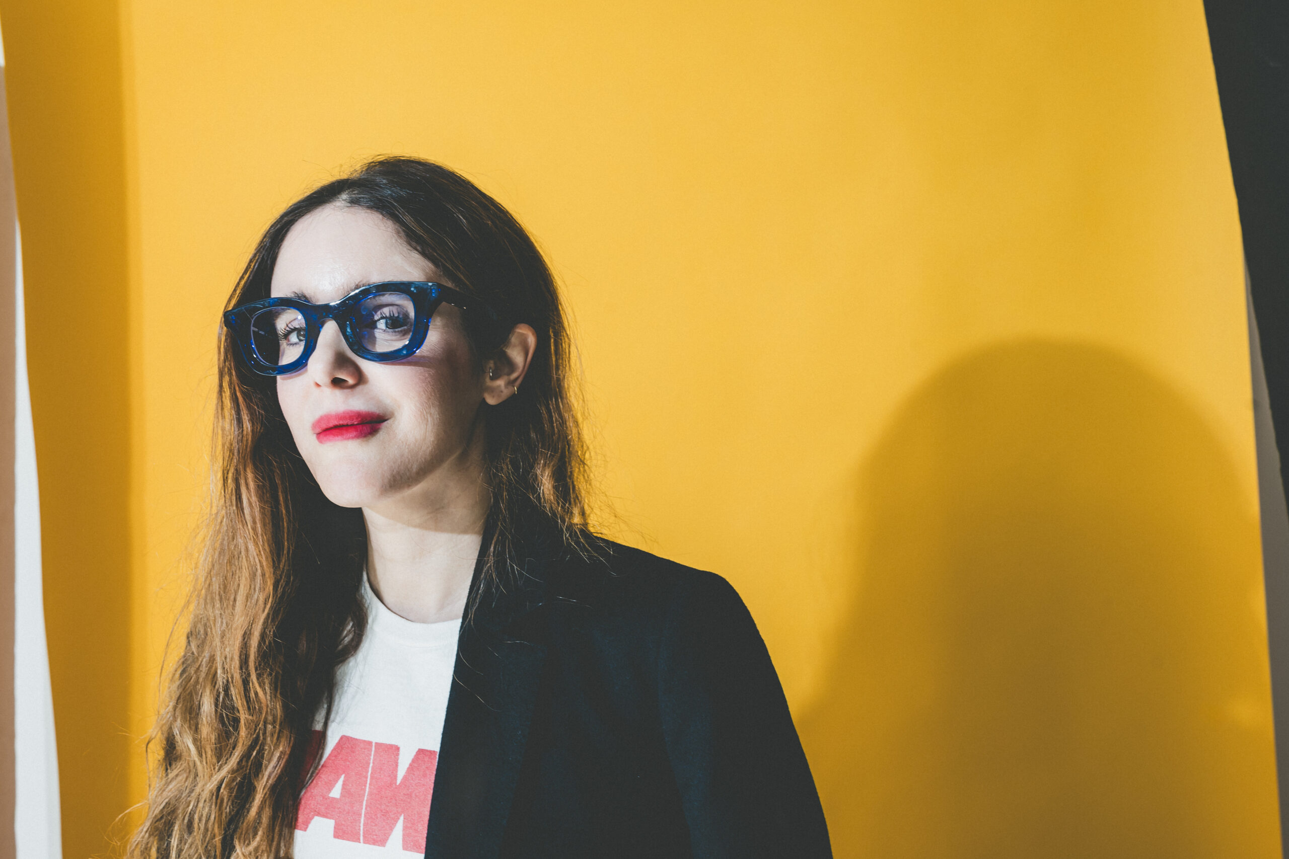 Woman wearing blue glasses and a black blazer stands against a yellow backdrop with partial white shirt visible.
