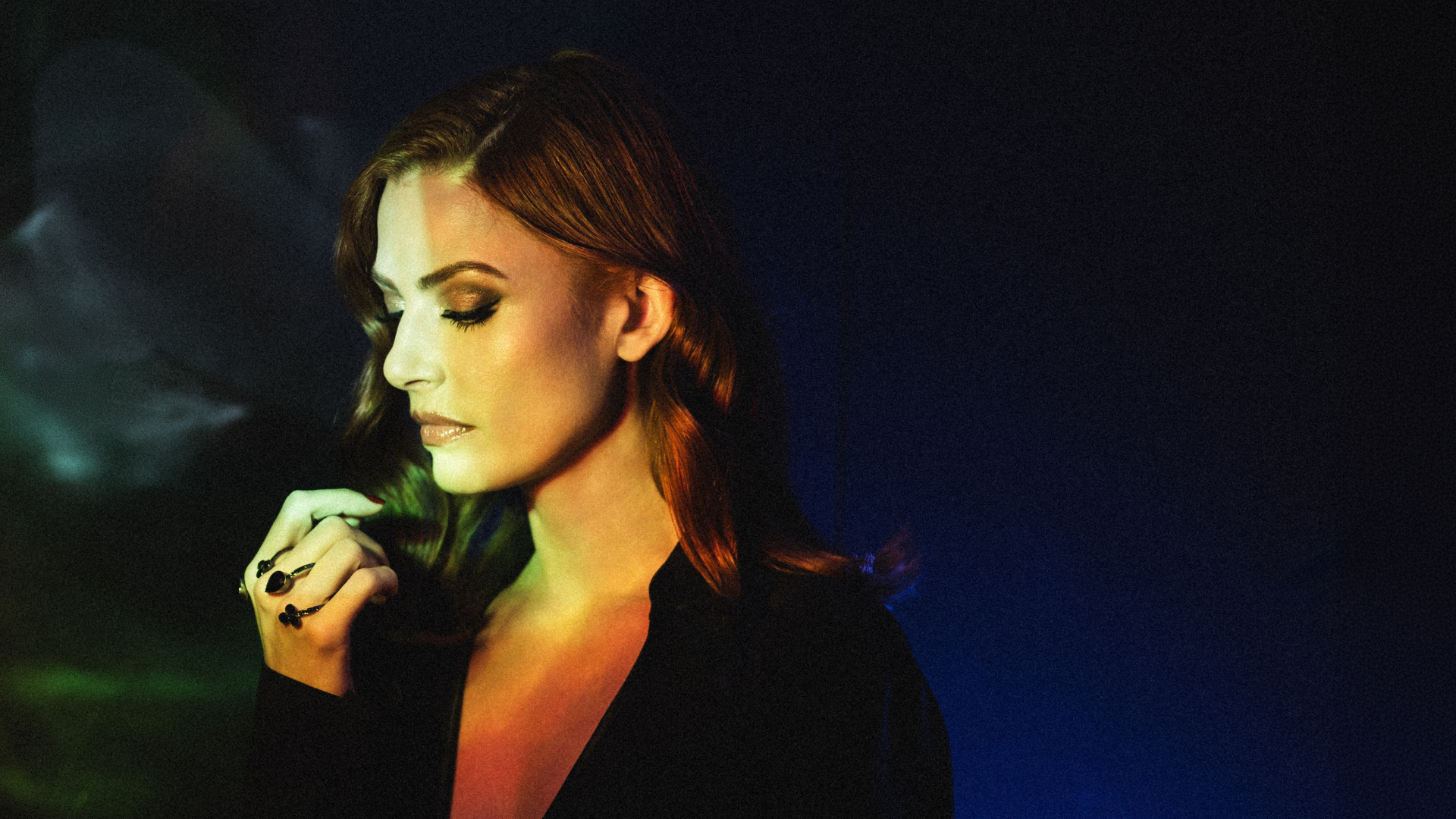 Portrait of a woman with smoky makeup and glossy lips, facing left under colorful stage lighting.