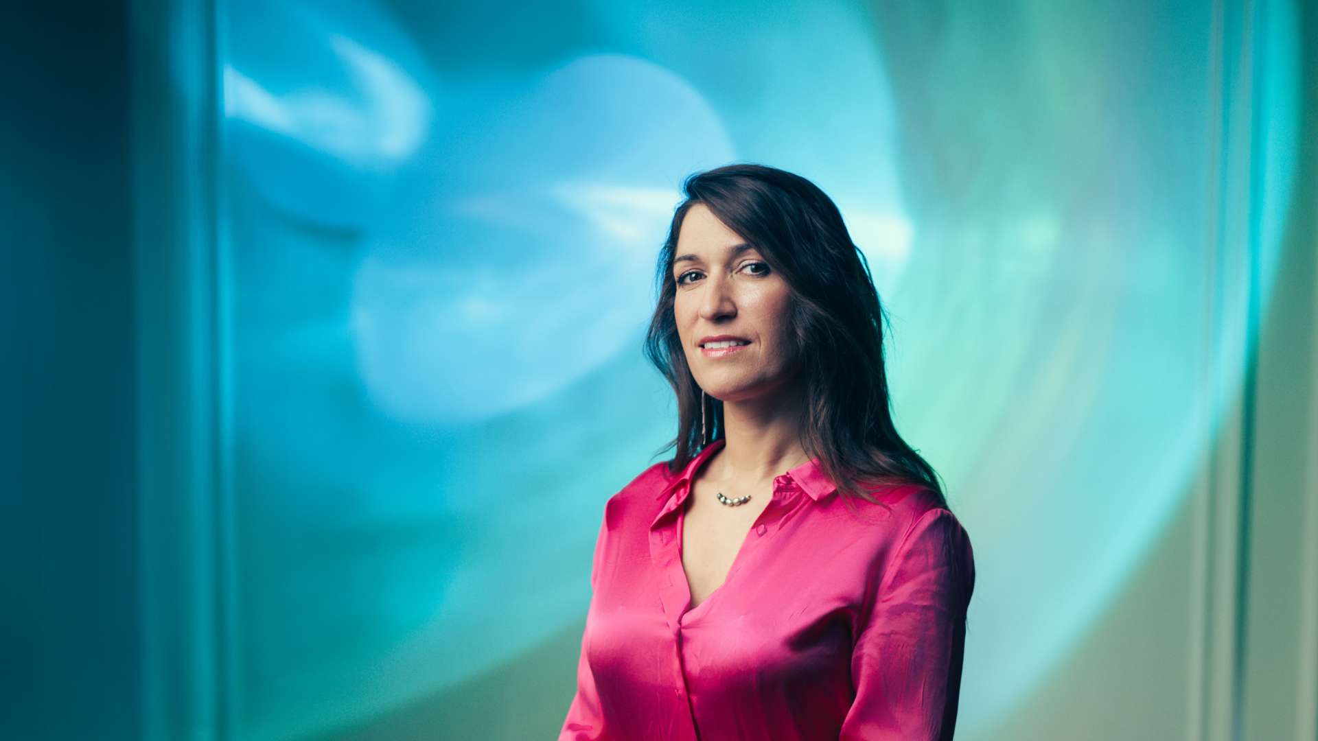 Portrait of a woman wearing a pink shirt, facing the camera, against a blue-green abstract background.