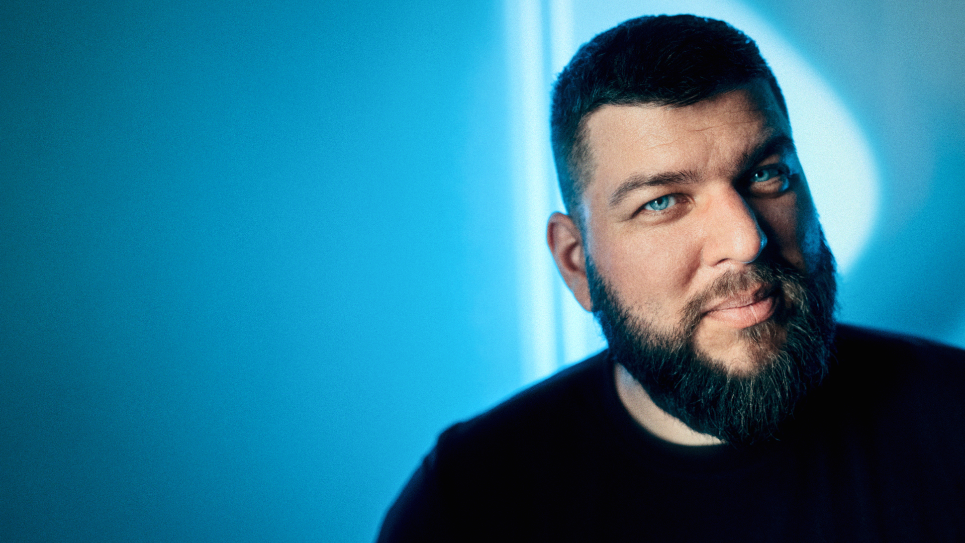 Close-up portrait of a bearded man looking at the camera with blue lighting on his face and background.