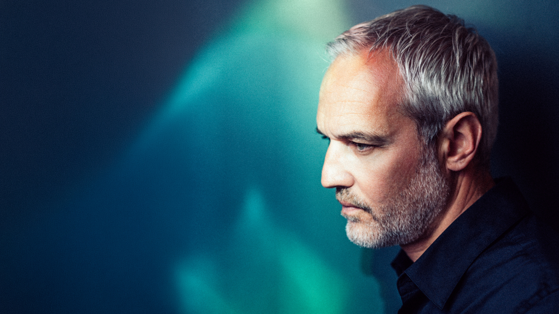 Side profile of a middle-aged man with gray hair and stubble against a teal-blue background.