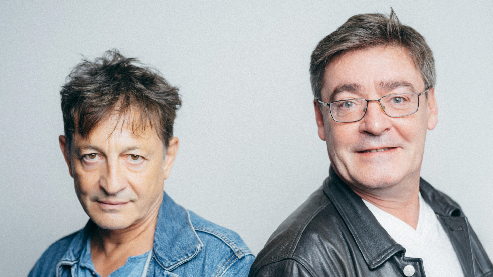 Two middle-aged men posing together against a gray backdrop, one in a denim jacket and the other in a leather jacket.